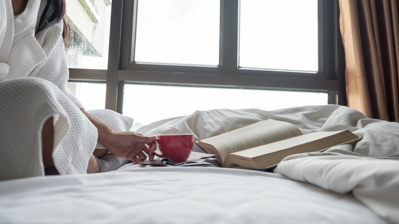 A woman in a gown reading a book on her bed with a cup of coffee